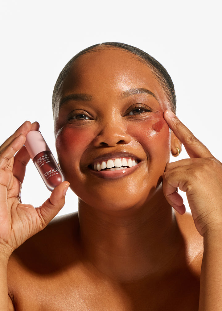 Model holding Annadea Power Walk Blush Serum while blending a rosy flush onto cheekbones, smiling with glowing skin