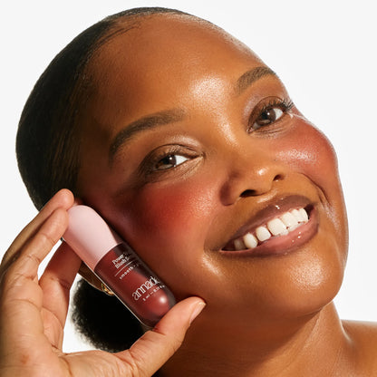 Smiling model with deep skin tone holding the Annadea Power Walk Blush Serum in Late Date against her cheek, showing a rich warm terracotta flush and luminous dewy glow from the smoothing and plumping blush serum
