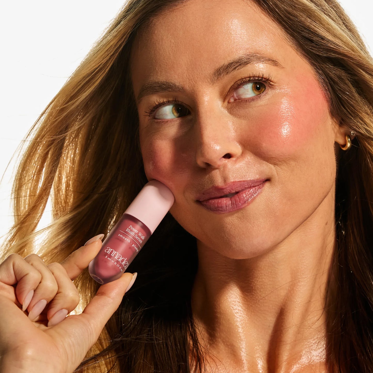 Model with light skin tone holding the Annadea Power Walk Blush Serum in Rose Trail against her cheek, showing a warm dusty rose flush and natural dewy glow from the smoothing and plumping blush serum