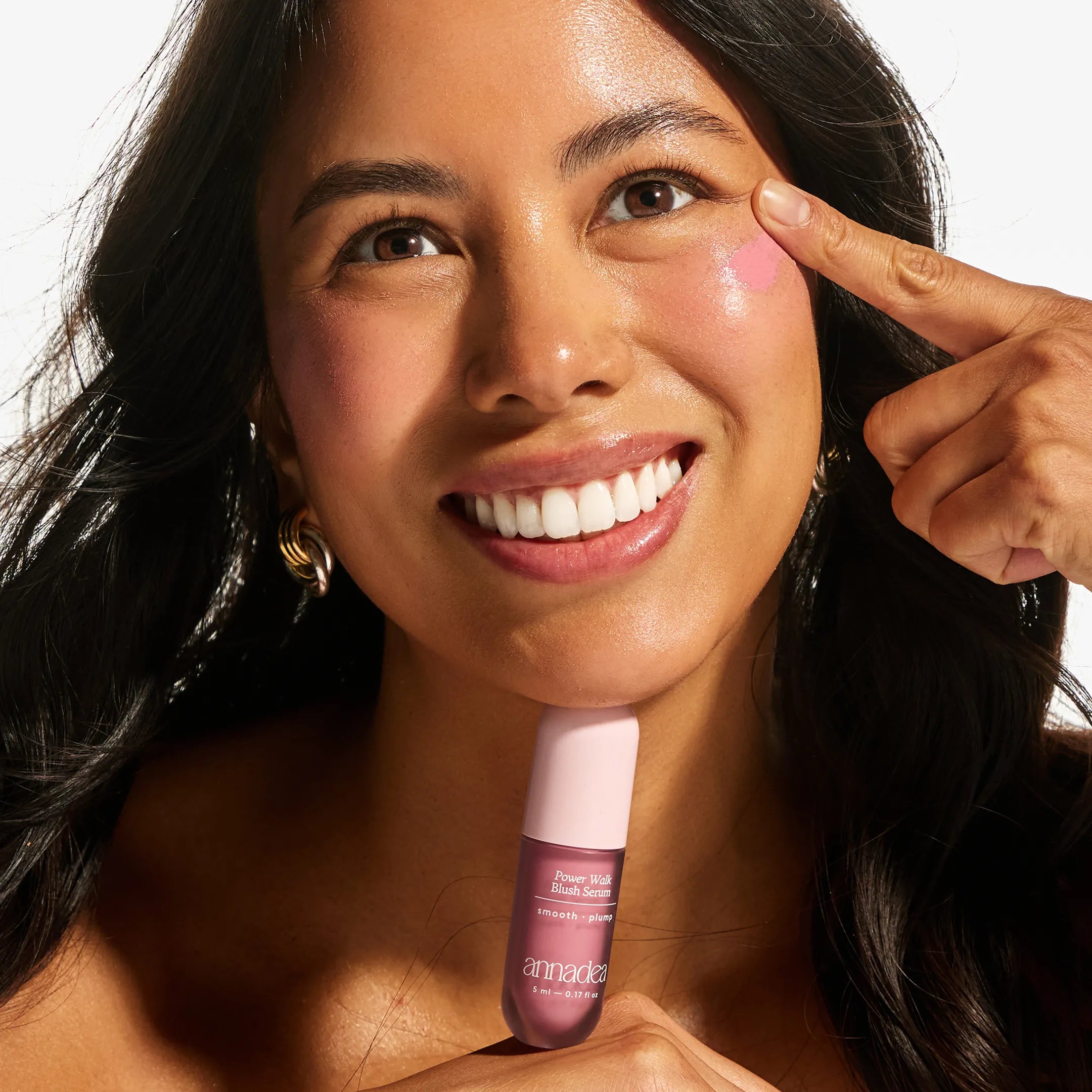 Smiling model with medium skin tone applying the Annadea Power Walk Blush Serum in Soft Step by dotting it onto her cheek, holding the bottle and demonstrating the buildable blush serum application technique