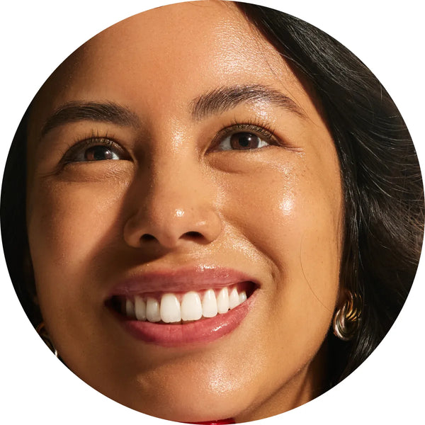 Close-up circular crop of a smiling model with medium skin tone showing luminous, dewy radiance from the Annadea To Dew Glow Serum, reflecting the serum's soft-focus lit-from-within glow