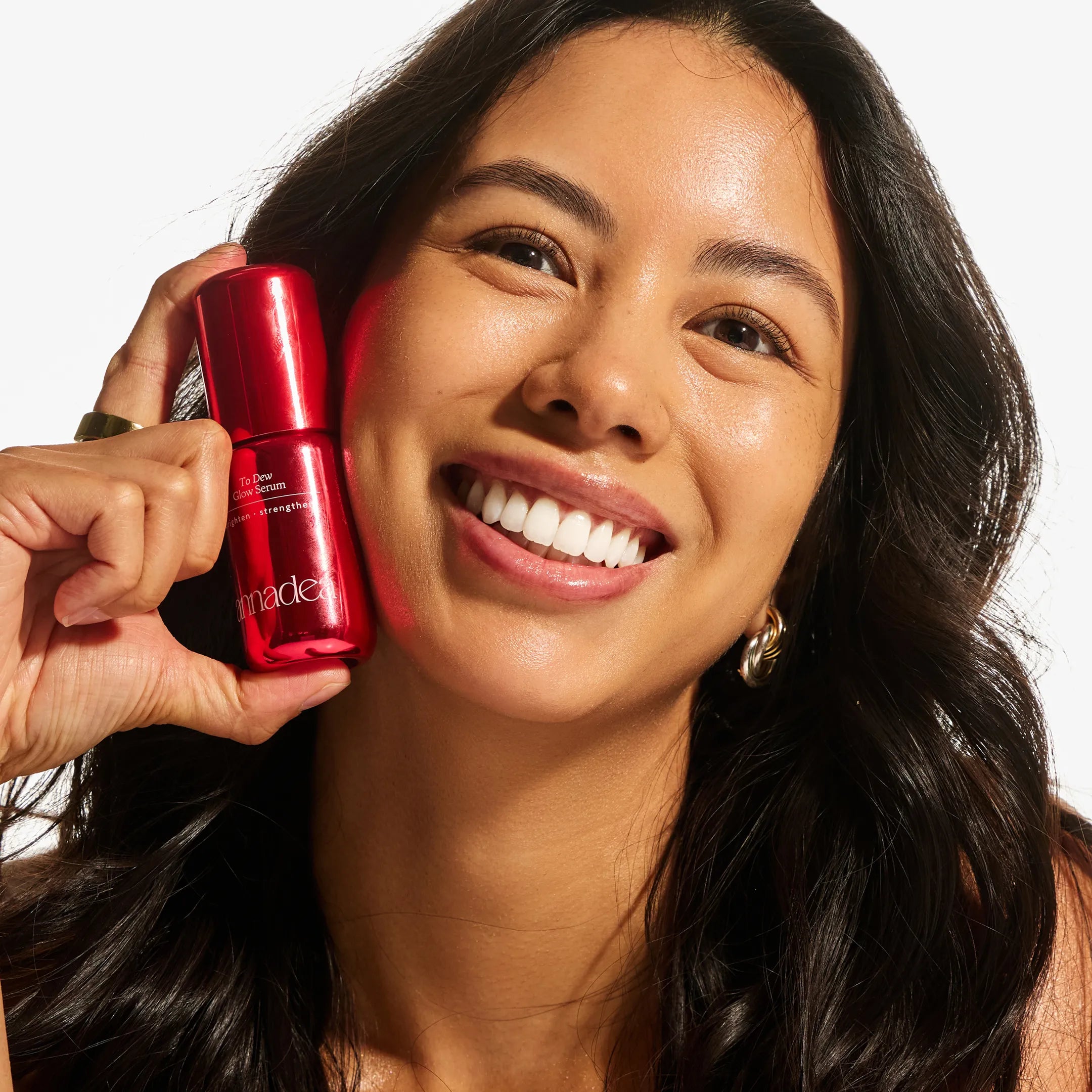 Smiling model with glowing, dewy skin holding the Annadea To Dew Glow Serum red bottle close to her cheek, showcasing the brightening and strengthening serum