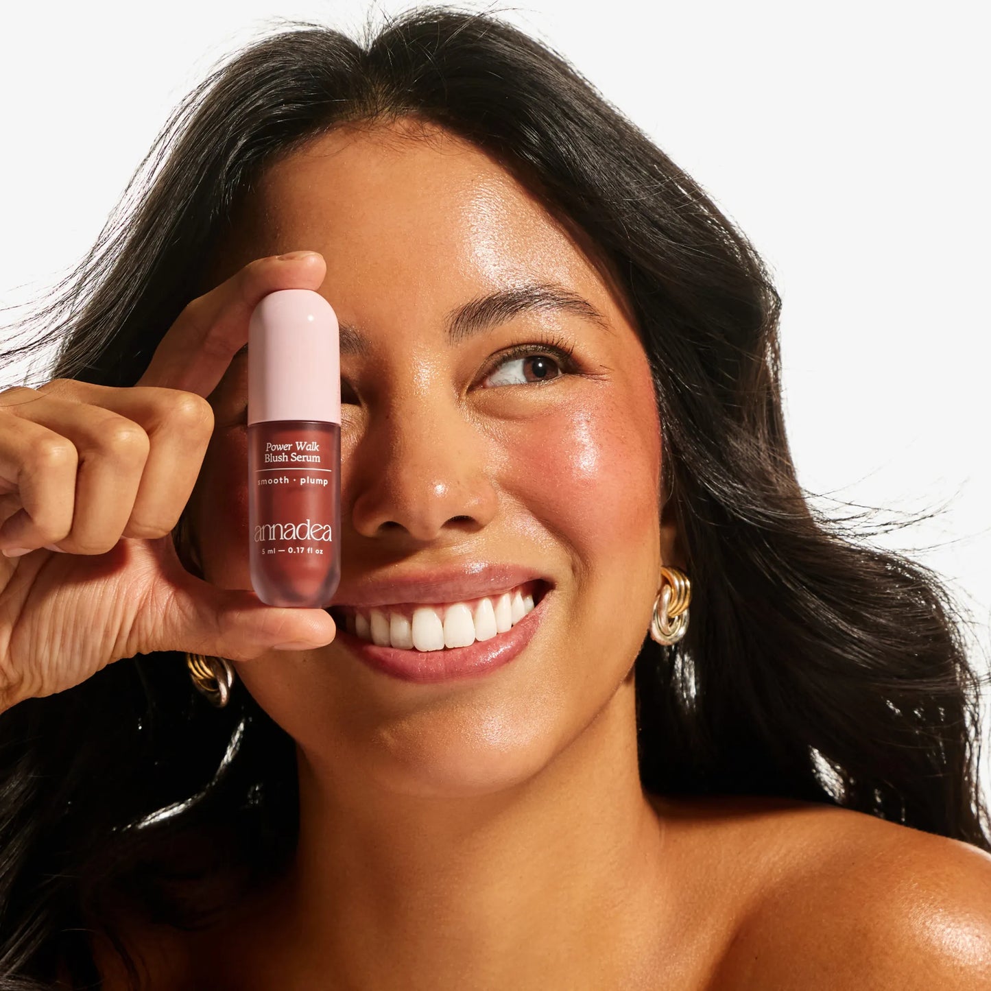 Smiling model with medium skin tone holding the Annadea Power Walk Blush Serum in Late Date in front of her face, showing a sun-kissed bronzed flush and radiant glow from the buildable blush serum