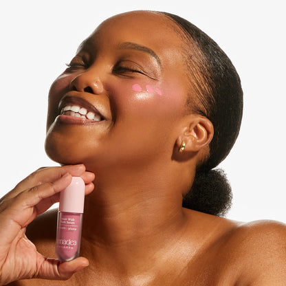 Laughing model with deep skin tone holding the Annadea Power Walk Blush Serum in Soft Step with baby pink serum dots visible on her cheek before blending, demonstrating the buildable application for a sheer natural flush