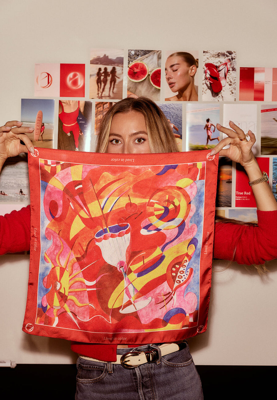 Annadea founder Olivia Adriance holding up the limited-edition Lived in Color silk scarf designed by artist Henriette Arcelin, featuring a vibrant abstract design in red, yellow, pink, and blue in front of the Annadea brand moodboard
