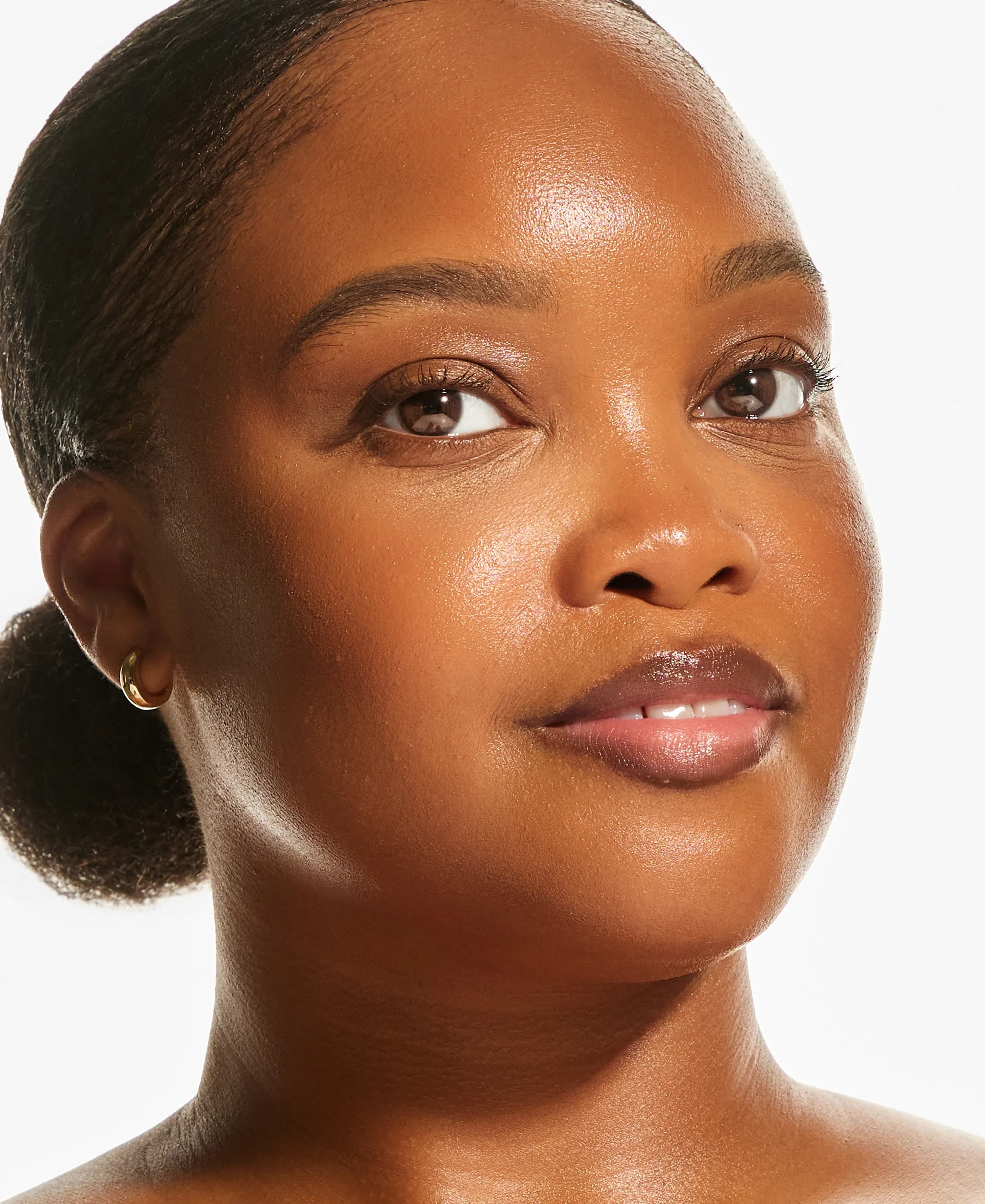 Model with deep skin tone wearing Annadea PTO Glow Bronzing Serum, demonstrating radiant, glowing complexion with small gold hoop earring against a white background