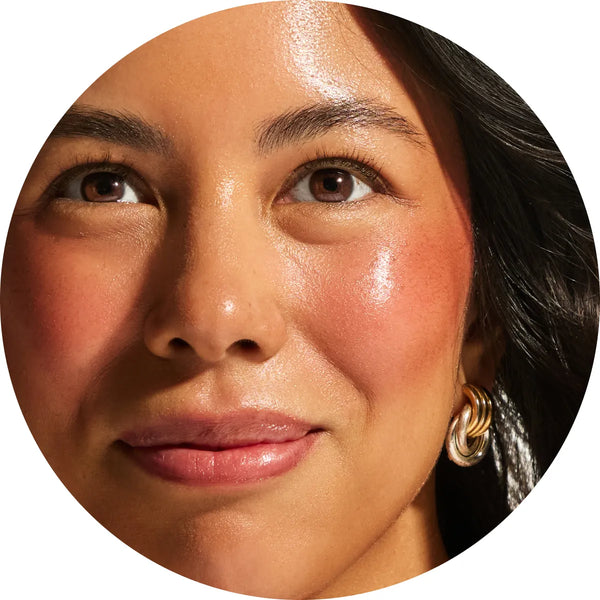 Close-up circular crop of a model with medium skin tone showing a warm bronzed terracotta flush across the cheeks from the Annadea Power Walk Blush Serum in Late Date, highlighting the serum's naturally flushed dewy finish and plumping results
