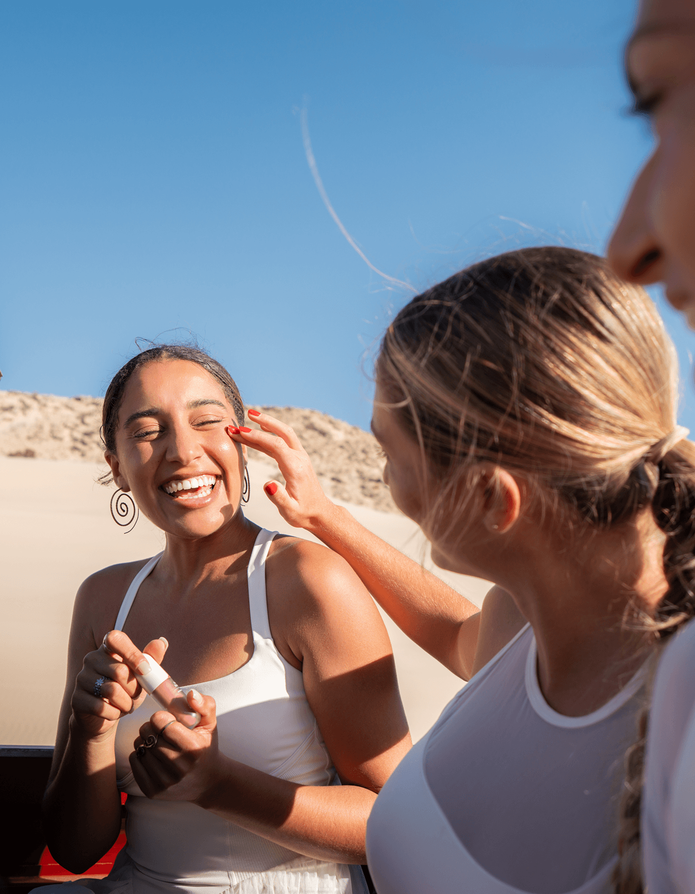 Two women outdoors on a sunny day, one applying Annadea Power Walk Blush Serum to the other's cheek against a blue sky and sand dunes backdrop, Annadea lifestyle campaign