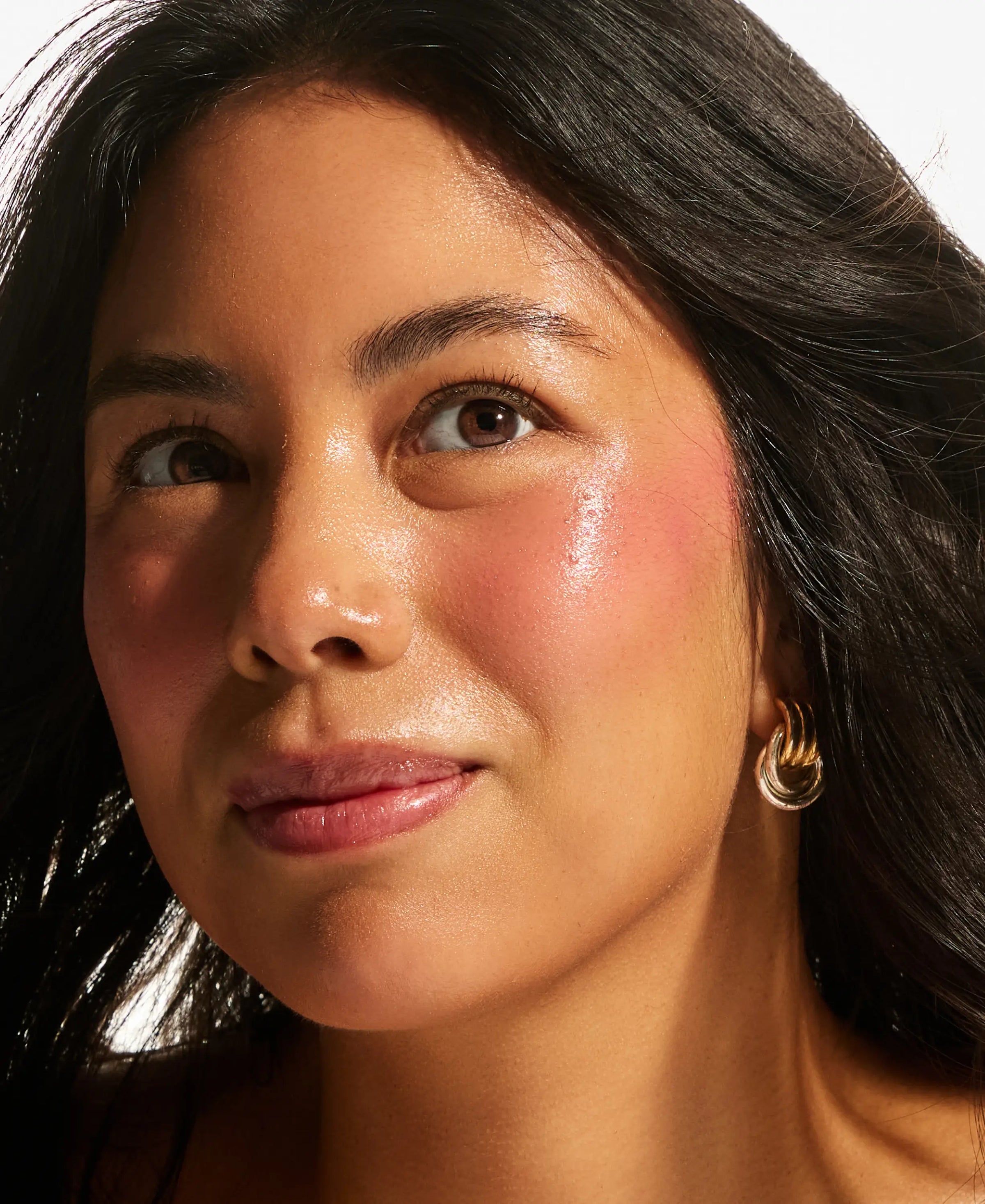 Model with medium skin tone wearing Annadea Power Walk Blush Serum, showing buildable rosy flush and luminous glow across cheekbones, with gold knot earring against a white background