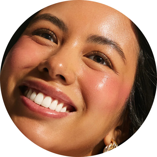 Close-up circular crop of a smiling model with medium skin tone showing a luminous rose flush across the cheeks from the Annadea Power Walk Blush Serum in Rose Trail, highlighting the serum's buildable natural rosiness and hydrating glow