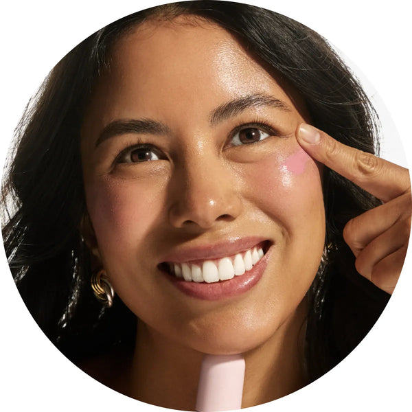 Close-up circular crop of a smiling model with medium skin tone pointing to a dot of Annadea Power Walk Blush Serum in Soft Step on her cheek before blending, demonstrating the serum application technique for a buildable baby pink flush