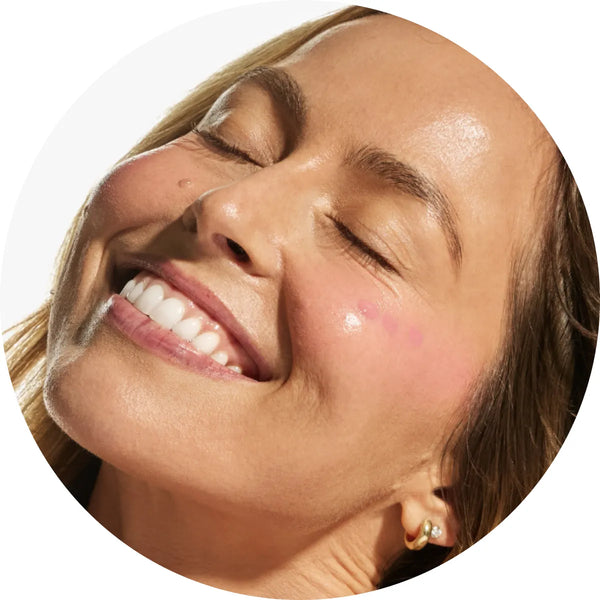 Close-up circular crop of a smiling model with light skin tone showing a soft sheer pink flush across the cheeks from the Annadea Power Walk Blush Serum in Soft Step, reflecting the serum's naturally flushed dewy finish and plumping results