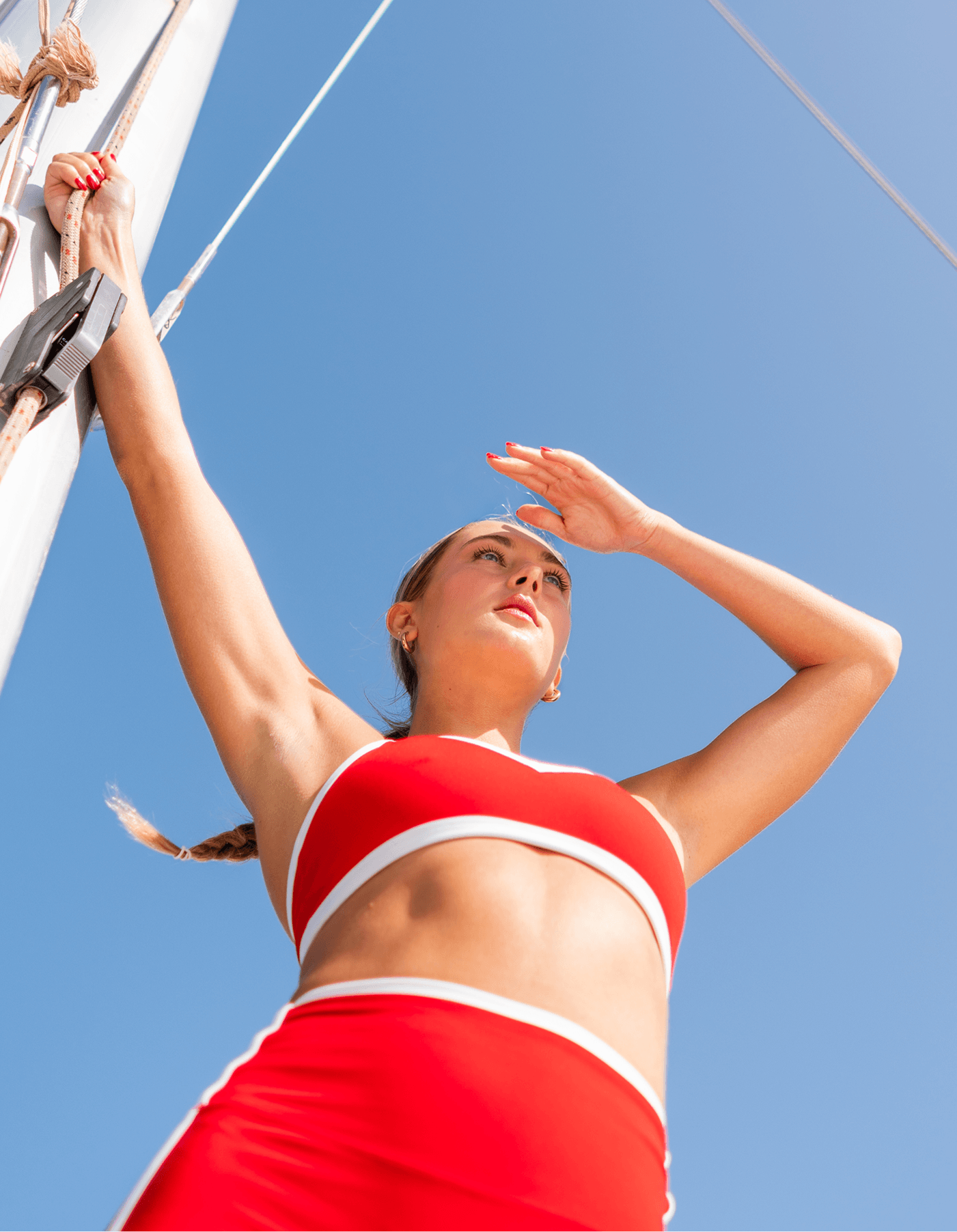 Woman in red athletic set on a sailboat shielding her eyes from the sun against a clear blue sky, Annadea To Dew Glow Serum lifestyle campaign