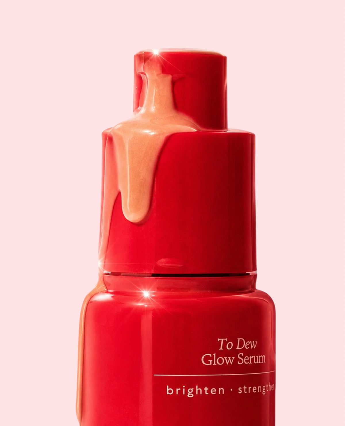  Close-up of the Annadea To Dew Glow Serum pump top with amber serum dripping down the red bottle against a soft pink background, showing the lightweight velvety serum texture